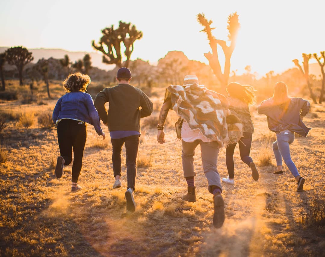 Group of 5 people running away from the camera in an arid-looking environment with the sun near the horizon