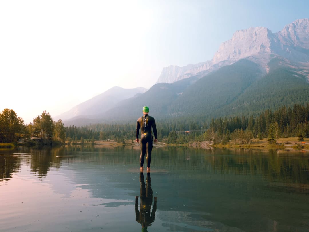 Man standing in a lake wearing a wetsuit, in the background are mountains.