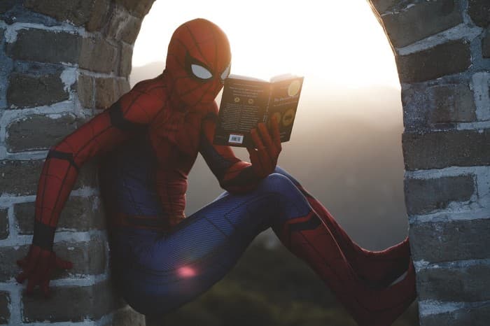 Someone wearing a Spiderman costume hanging on to an archway and holding a book