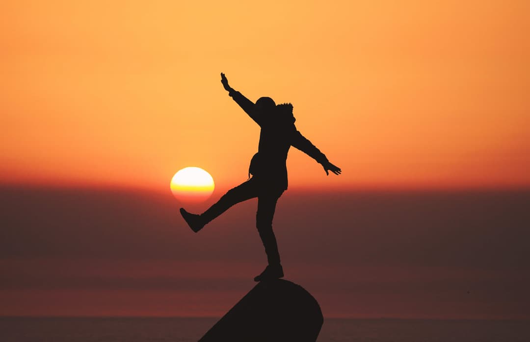 Photo of person standing in front of a rock, with the sun setting in the distant background