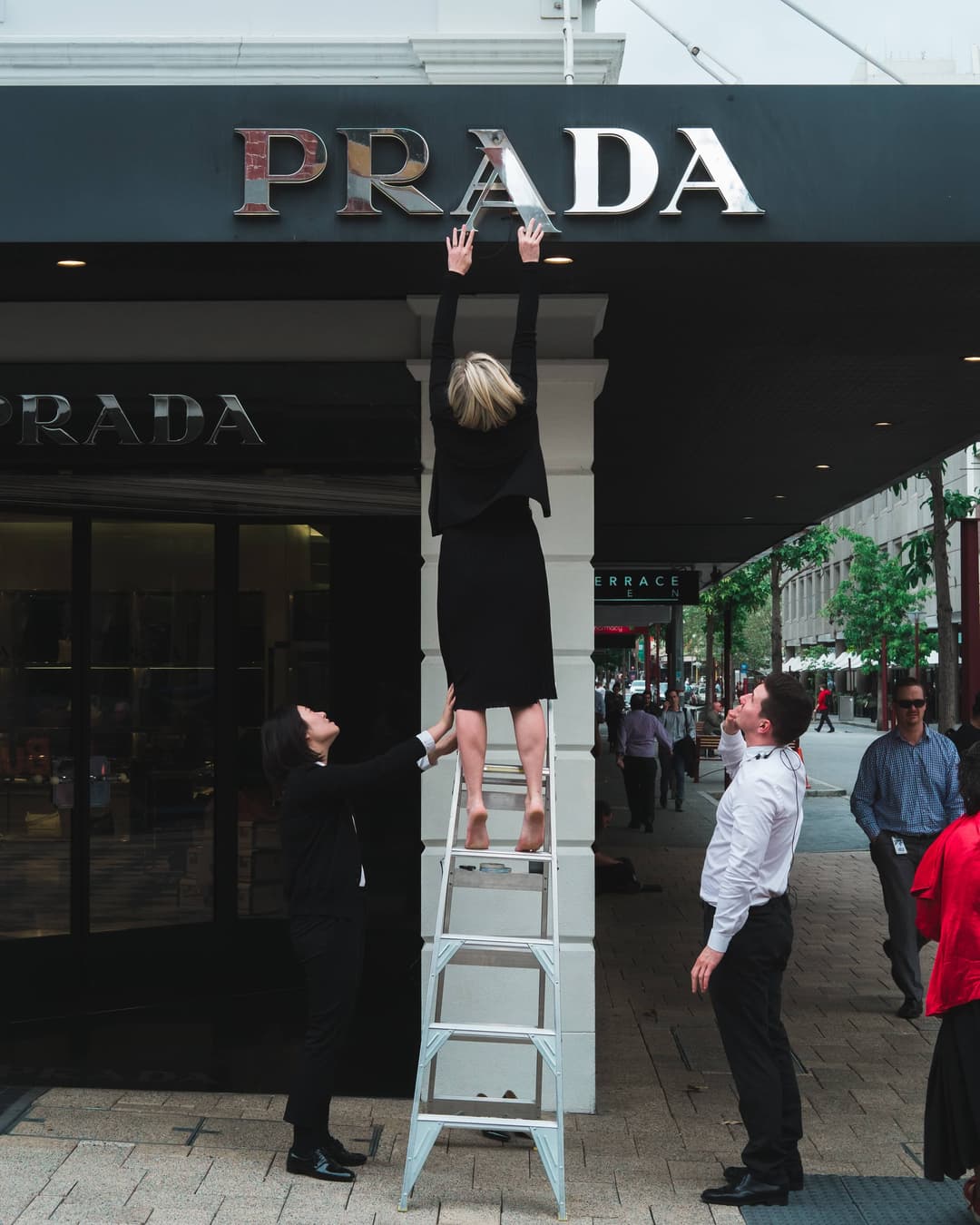 A woman standing on a ladder fixing up the alignment of the centre 'A' in a 'Prada' logo on a storefront, with two people watching her on the side.